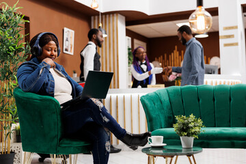 Young African American woman traveler sitting with laptop in hotel lobby using free wireless...