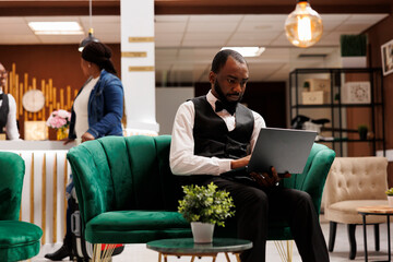 Focused African American man hotel manager sitting in lobby working on laptop, looking at computer...