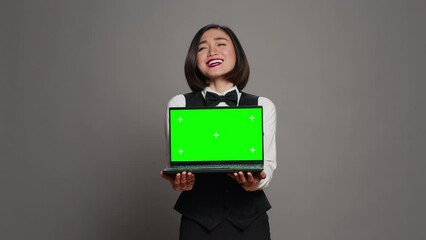Receptionist showing greenscreen display on personal laptop, presenting isolated copyspace mockup on screen. Asian hotel employee holding pc with chromakey mockup template. Camera A.