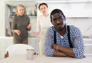 Sad middle-aged man sitting at the kitchen table with his back to old and middle-aged women...