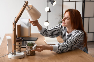 Pretty young woman changing light bulb in desk lamp at home - obrazy, fototapety, plakaty
