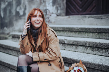 A happy stylish ginger lady sitting on old stairs and taking on a phone