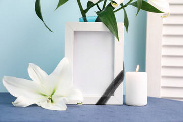 Blank funeral frame, burning candle and vase with lily flowers on table near color wall, closeup - obrazy, fototapety, plakaty