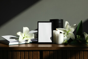 Blank funeral frame, burning candles, mortuary urn and lily flowers on wooden cabinet against grey...