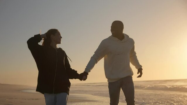 Casually dressed loving young couple running hand in hand through waves on shoreline watching beautiful sunrise morning over beach and sea in South Africa - shot in slow motion