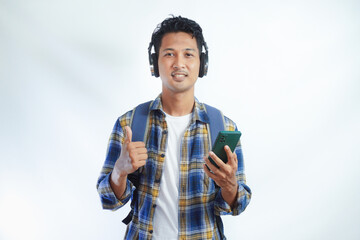 Smiling young Asian man in casual shirt and backpack with mobile phone and wireless headphones, showing thumbs up isolated on white background. People emotions lifestyle concept