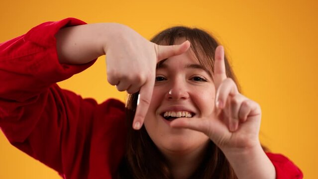 Portrait of woman looking at camera through finger frame isolated on yellow background with copy space