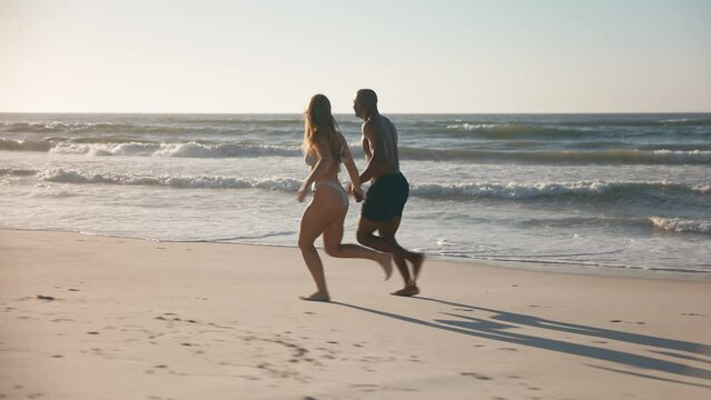 Camera tracks full length shot of loving young couple in swimwear holding hands and running along beach in South Africa - shot in slow motion