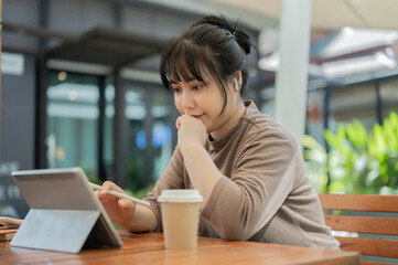 A focused Asian woman is working remotely at a cafe, wearing earbuds and using her digital tablet.