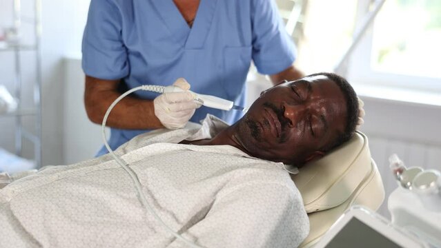 Male cosmetologist performs skin care and maintenance procedure for African-American man client. Specialist uses scriber nozzle to remove dead particles and peel-off clarifying