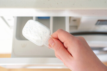 Female hand pours a scoop of powder into washing machine, top view.