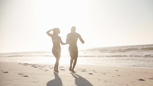 Camera tracks full length shot of loving young couple in swimwear holding hands and running along beach in South Africa shot against sun - shot in slow motion