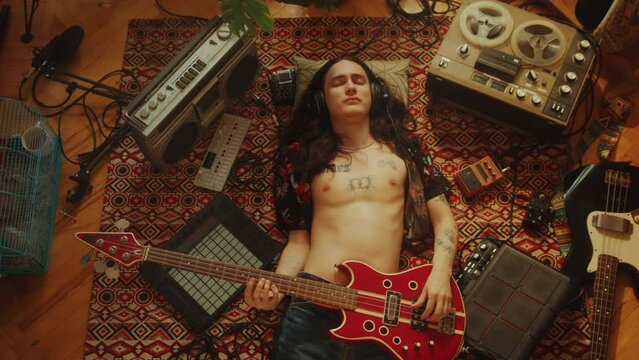 Young relaxed musician in headphones and open shirt playing electric guitar with eyes closed while lying down on floor among retro tape recorders and audio devices. Directly above view