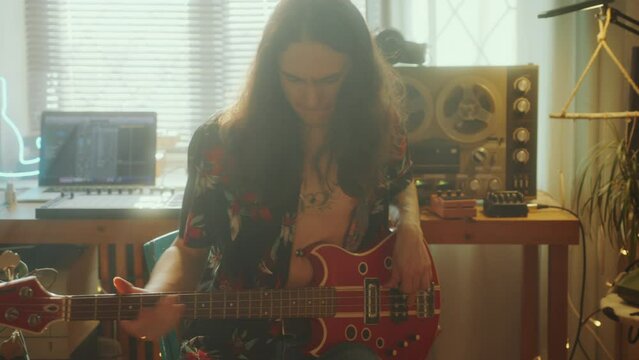 Young long-haired rock musician playing electric guitar in front of desk with laptop and vintage reel-to-reel tape player in sunlit home studio