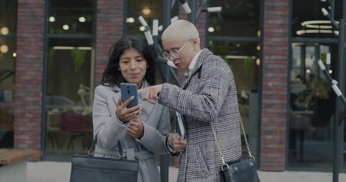 Slow motion portrait of cheerful businesswomen discussing online content on smartphone screen talking outdoors in modern city street