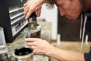 Young hispanic man pouring thinner into a cup of black paint on a workshop countertop. Real people at work.