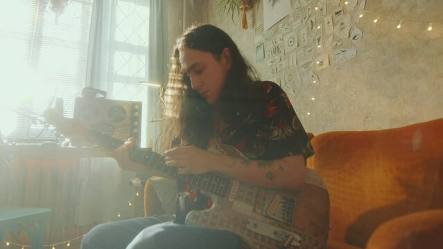 Young guy playing electric guitar while sitting on couch in a sunlit room with creatively designed interior, cozy string lights and retro tape recorders. Zoom shot