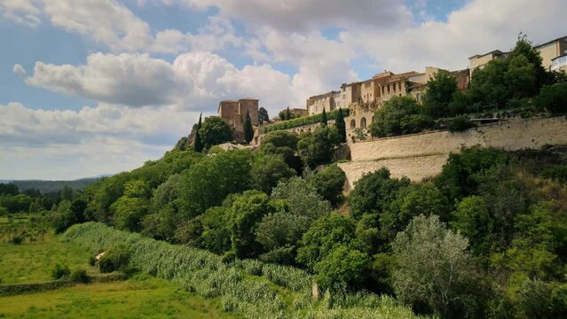 Drone ascends forested hillside above farmland fields to historic town archway walls of Lauris France