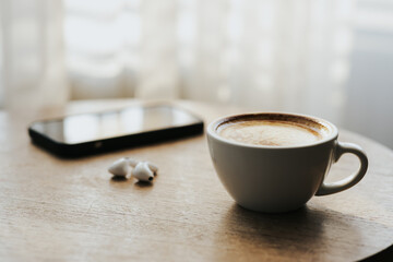 A cup of hot latte and a cell phone, headphones on a sunny wooden table by a window with white...