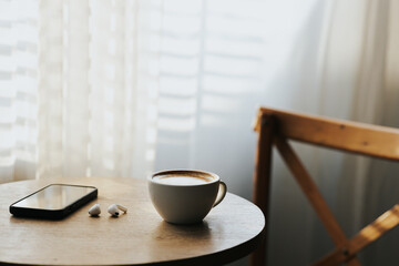 A cup of hot latte and a cell phone, headphones on a sunny wooden table by a window with white curtains. Vintage tones. Free space to create a message.