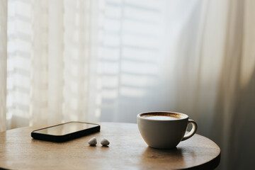 A cup of hot latte and a cell phone, headphones on a sunny wooden table by a window with white curtains. Vintage tones. Free space to create a message.