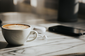 A cup of hot latte and a cell phone, headphones on a sunny wooden table by a window with white curtains. Vintage tones. Free space to create a message.