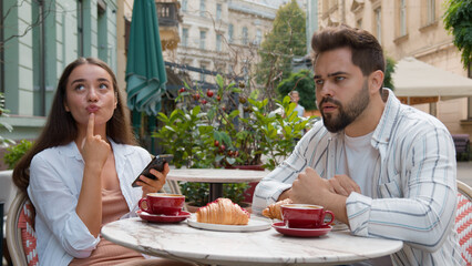 European young couple in city cafe woman smiling laughter emotional texting smartphone ignore man fixed gaze distant frustrated relationship problem indifference outdoors breakfast internet addiction