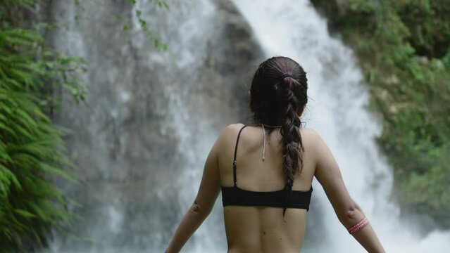 Beautiful woman in black bikin top stares out to gorgeous tropical waterfall in forest