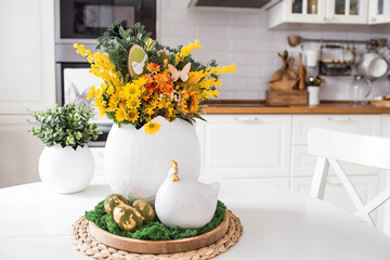 A spring bouquet in an egg, an Easter chicken and eggs with a golden pattern on the table. In the background is a white Scandinavian-style kitchen. Easter decor in the house.