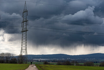 Strommast einer Überlandleitung vor dramatischen Himmel mit einer abregnenden, grauen Gewitterwolke und Mittelgebirge am Horizont