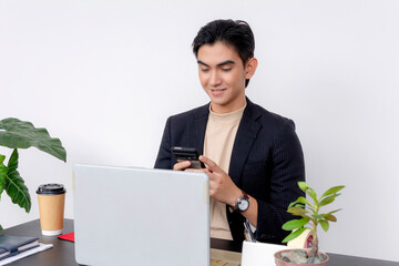Young Asian businessman smiling or amused while checking his cellphone, with a coffee cup and computer on his desk.
