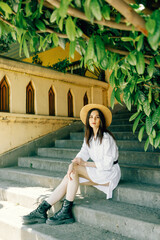 a cute girl in a hat is sitting on the stairs of the old town. Brunette in a white dress walks the street - 745589557