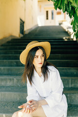 a cute girl in a hat is sitting on the stairs of the old town. Brunette in a white dress walks the street - 745589904
