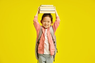 Cute chinese schoolgirl holding books on head over yellow isolated background. First grader kid...