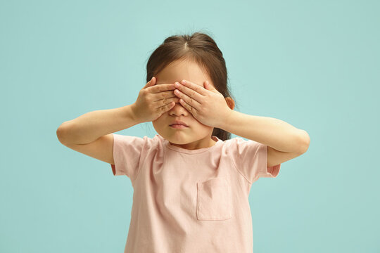 Kid girl with serious face covering her eyes with hands from scared or playing peekaboo standing over blue isolated