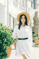 A cute girl in a hat walks along a tourist street among cafes. a young brunette in a white dress enjoys the city - 745591902