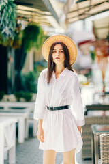 A cute girl in a hat walks along a tourist street among cafes. a young brunette in a white dress enjoys the city - 745592720