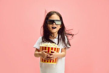 Joyful and Surprised Child Girl in 3D Glasses Holding Popcorn Bucket Having Flowing Hair Standing...
