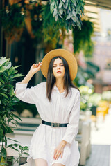 A cute girl in a hat walks along a tourist street among cafes. a young brunette in a white dress enjoys the city - 745592973