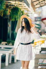 A cute girl in a hat walks along a tourist street among cafes. a young brunette in a white dress enjoys the city - 745593593