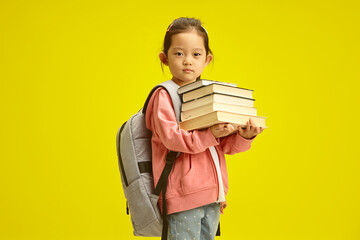 Happy cute industrious asian ethnicity child girl holding keeps a lot of books, and have backpack on shoulders, sweet looking to camera standing over yellow isolated background with a free copy space.