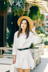 A cute girl in a hat walks along a tourist street among cafes. a young brunette in a white dress enjoys the city - 745593983