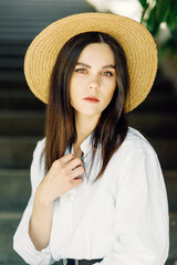 A close-up portrait of a young pretty fashionista posing in a European city. Cute brunette in a white shirt with a hat looks at the camera - 745594347