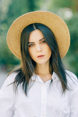 A close-up portrait of a young pretty fashionista posing in a European city. Cute brunette in a white shirt with a hat looks at the camera - 745594597