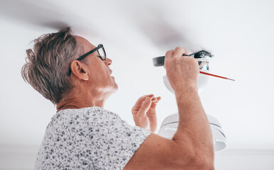 Mature experienced electrician on a ladder installing a paddle fan on the ceiling in the living room at home in preparation for the upcoming summer - obrazy, fototapety, plakaty