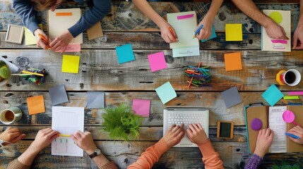 Close-up view of several hands collaboratively working on a project with colorful sticky notes, papers, and digital devices on a wooden table