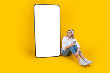 Mobile app mock up, full body length young blonde girl sit near huge smartphone with empty blank screen for mobile app mock up. Using phone, looking white huge display. Yellow background. Copy space.