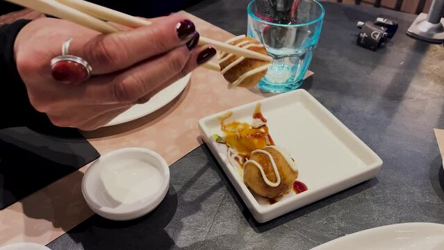 We see female hands with chopsticks taking some battered octopus balls, and soaking them in some sauces. With a few twists, she appears with painted nails in an Asian restaurant.