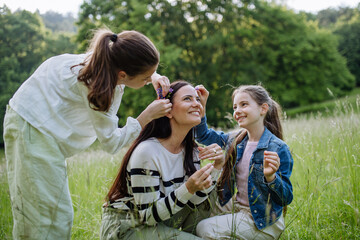 Fototapeta premium Beautiful mother with daughters, picking flowers, putting in hair, sitting in grass at meadow. Concept of Mother's Day and maternal love.