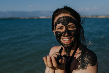 Woman applies healing mud to her face and body. Natural healing mud in Croatia on the beach. - obrazy, fototapety, plakaty